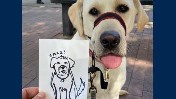 A hand holds up a cute drawing of K9 Cali while K9 Cali wags her tongue in the background.