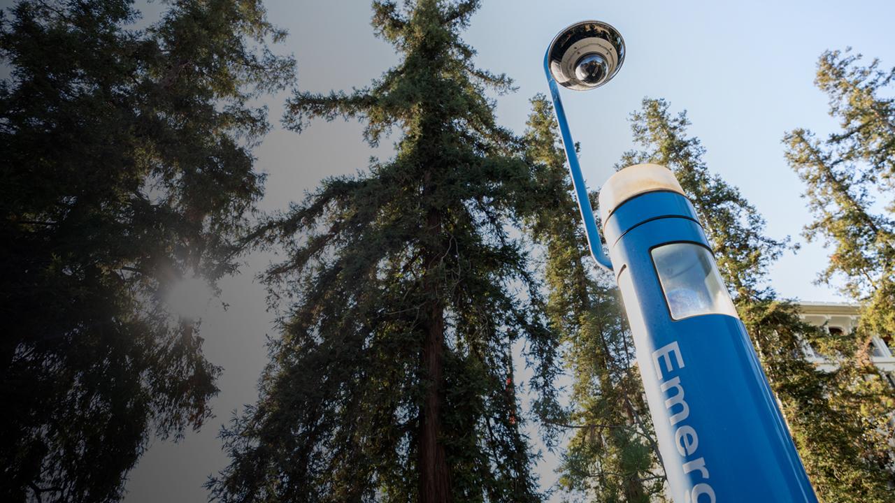 Emergency call box on campus surrounded by trees in the Arboretum and Mrak Hall in the background.