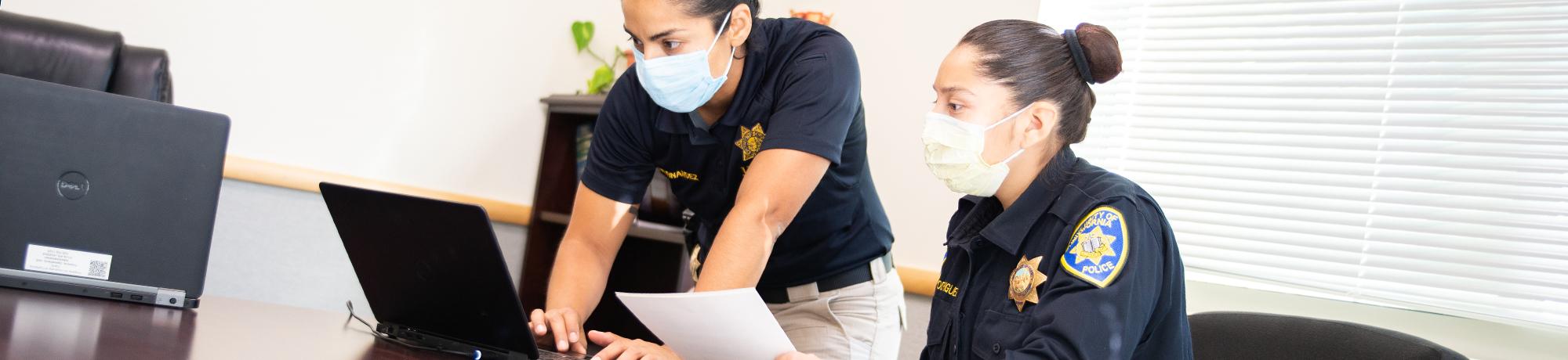Two female officers with face covers enter information onto a computer in a conference room