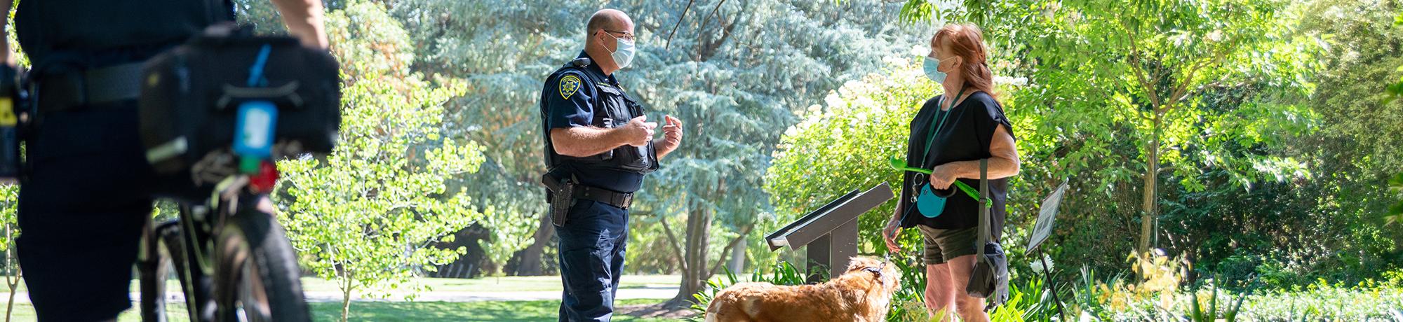 Officer and visitor with her dog talk in the arboretum while wearing face coverings