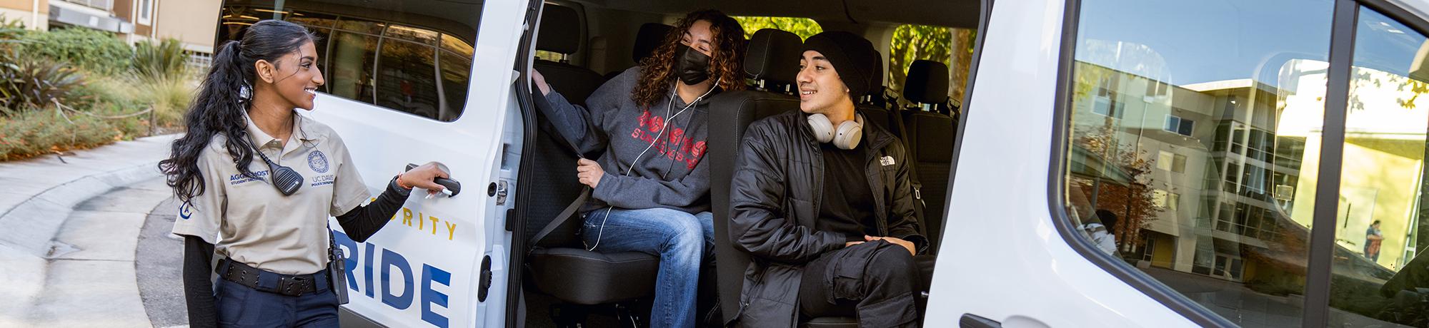 Aggie Host smiling and opening door of a Safe Ride van with two happy students sitting down inside.