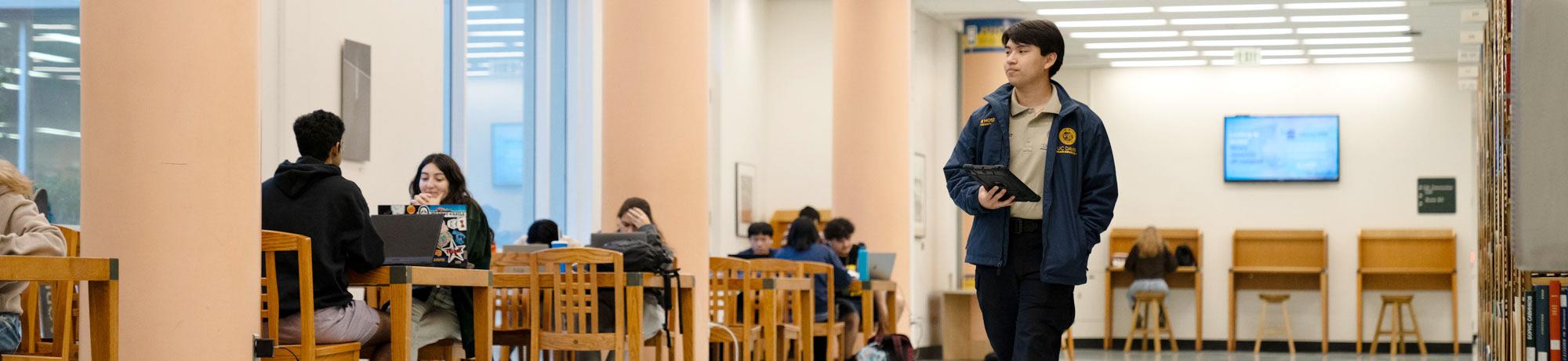 Aggie Host patrolling inside the UC Davis Library while students study in the background